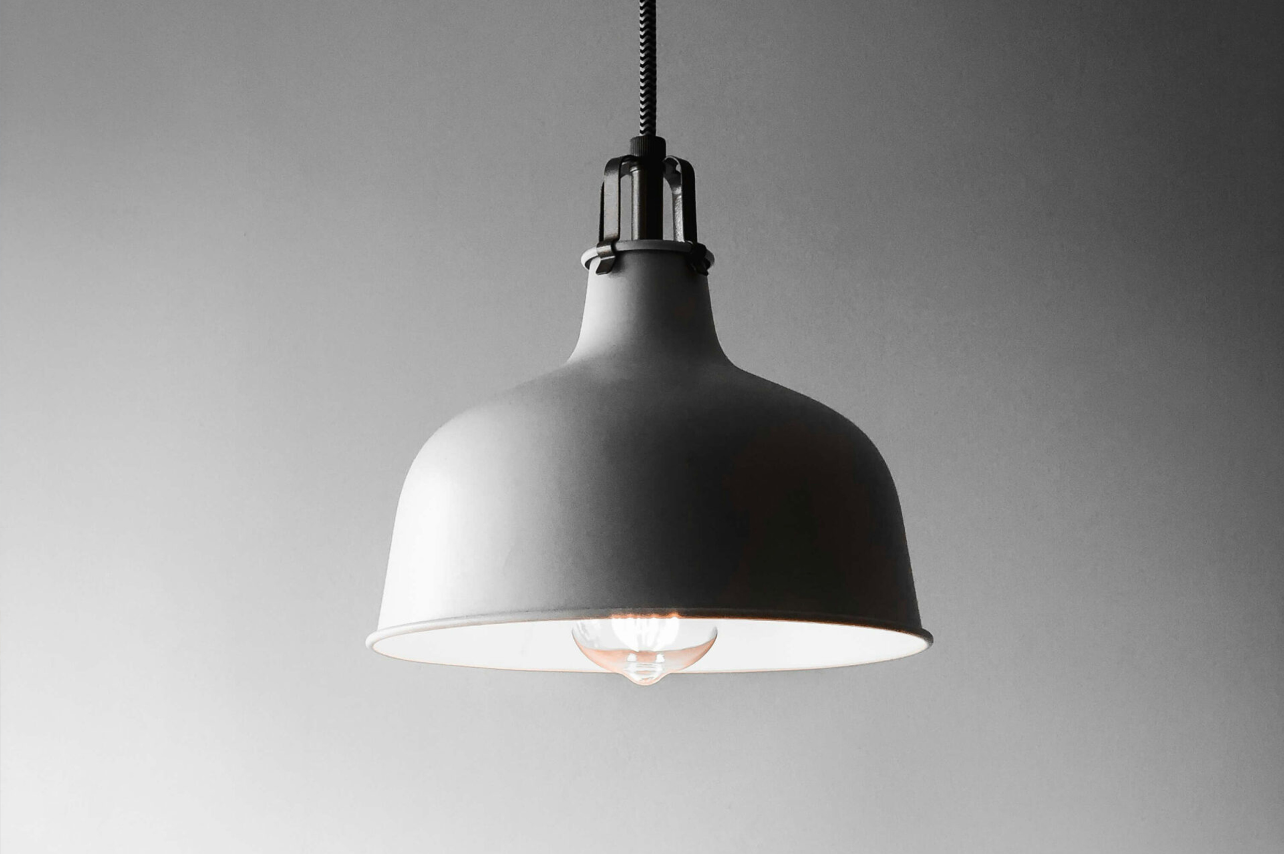 A single industrial-style pendant light fixture is hanging from the ceiling and is turned on, casting a soft glow against a plain gray background.
