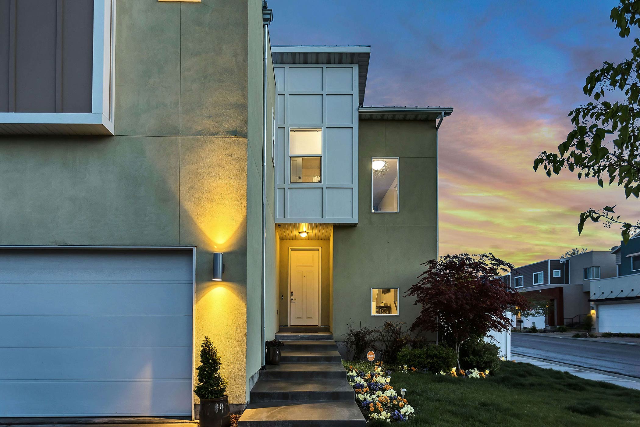 Modern two-story house with exterior lights on, a white garage door, landscaped front yard, and sunset sky in the background.