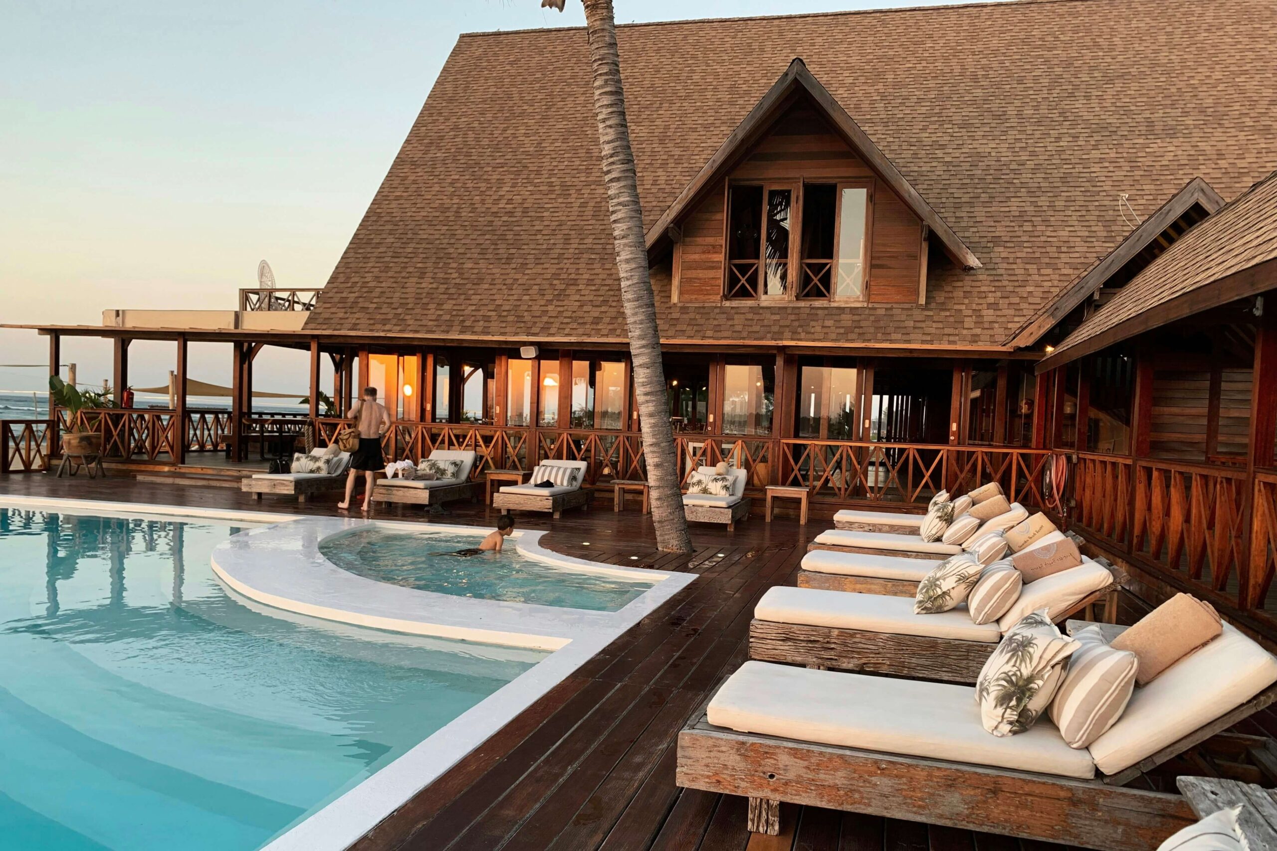 A wooden resort building with large windows, lounge chairs, and a pool on a deck at sunset. One person stands by the railing and another is near the water.