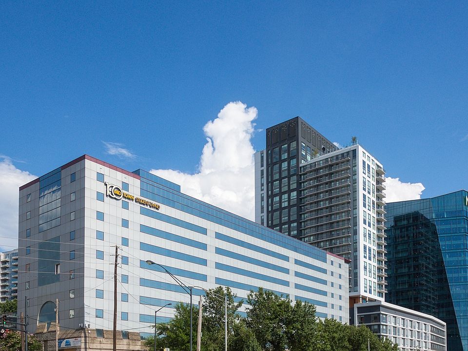 A multi-story office building with a "10th Anniversary" sign stands in front of modern high-rise buildings under a clear blue sky with clouds. Trees and streetlights are visible in the foreground.