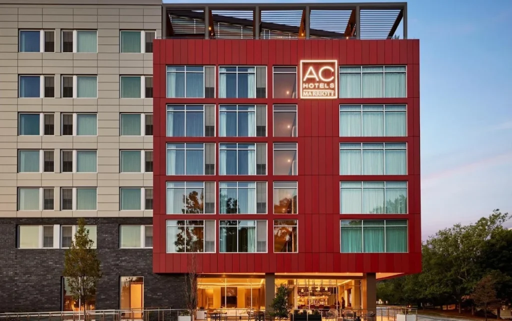 Exterior view of an AC Hotels by Marriott building with large windows, a red facade, and illuminated hotel signage at dusk.
