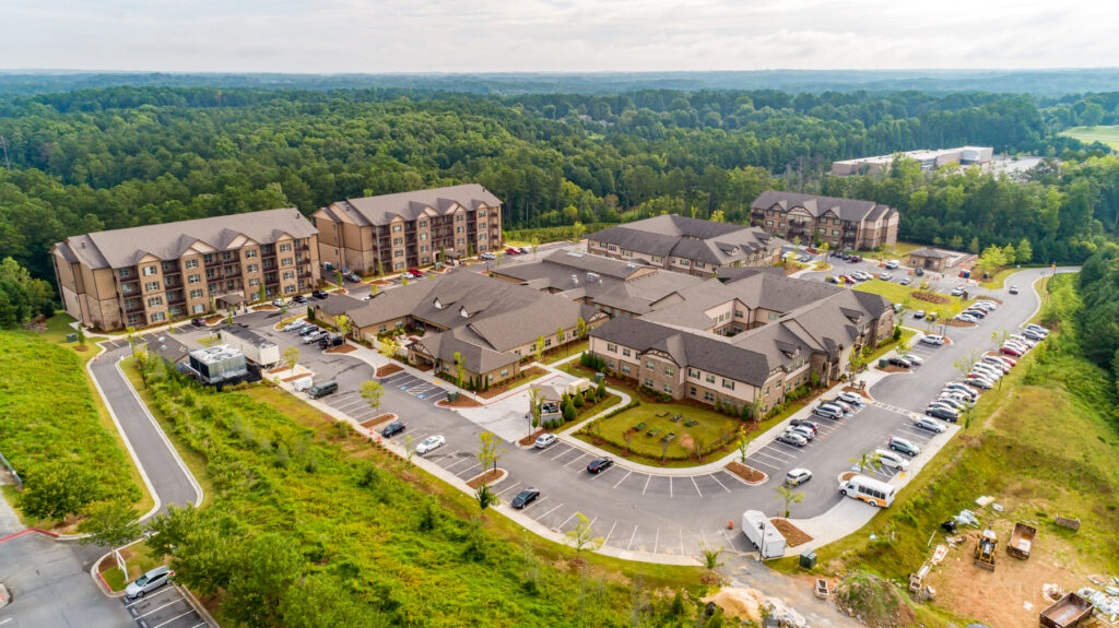 Aerial view of a large residential complex with multiple buildings, parked cars, landscaped grounds, and surrounding forested area.