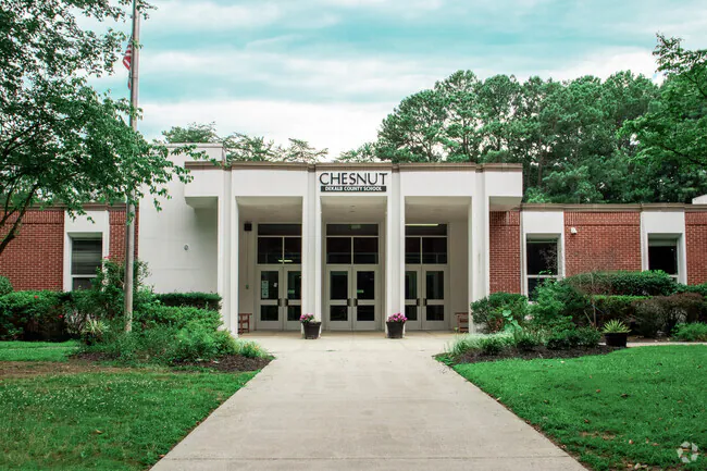 Front entrance of Chesnut Elementary School, a single-story brick and white building with a sign above the doors, surrounded by trees and landscaped bushes.