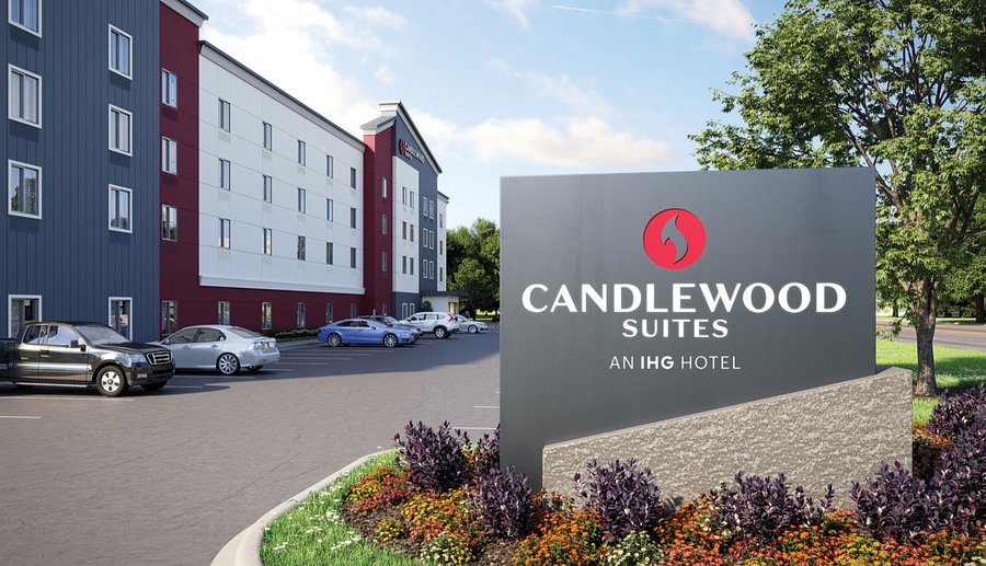 Candlewood Suites hotel exterior with parking lot, several parked cars, and large sign displaying the hotel name and logo in front of landscaped plants.