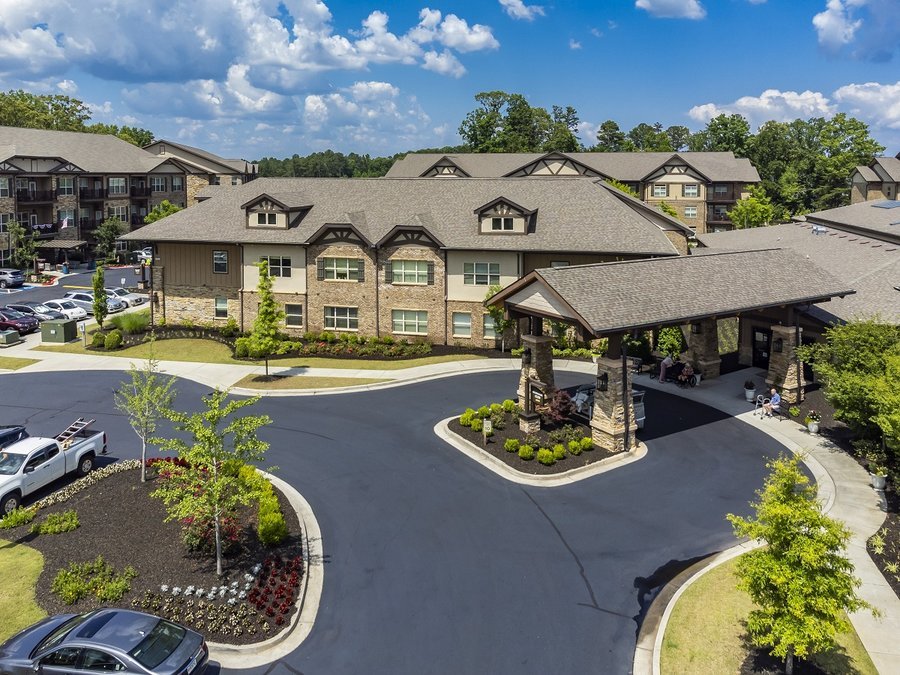 Aerial view of a large, modern assisted living facility with stone and brick exterior, landscaped grounds, and multiple parked vehicles.