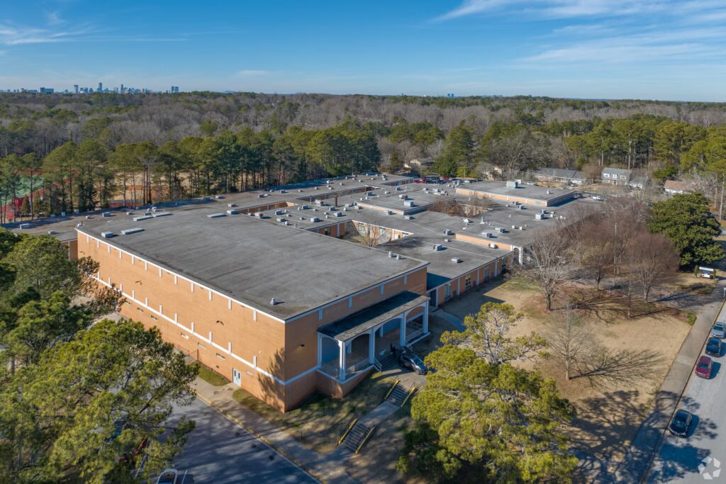 Aerial view of a large, rectangular brick building surrounded by trees and parking areas, with a city skyline visible in the distant background.