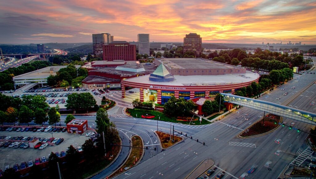 Aerial view of a convention center with a curved facade, adjacent parking lots, nearby office buildings, and intersecting roads at sunset.