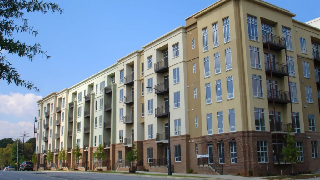 Four-story modern apartment building with large windows, small balconies, and a brick and stucco exterior, situated along a quiet street under a blue sky.