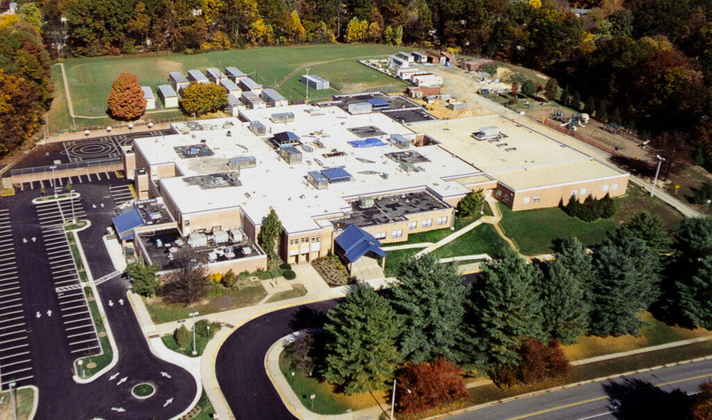 Aerial view of a large, flat-roofed school building with surrounding parking lots, sports fields, portable classrooms, and nearby wooded areas.