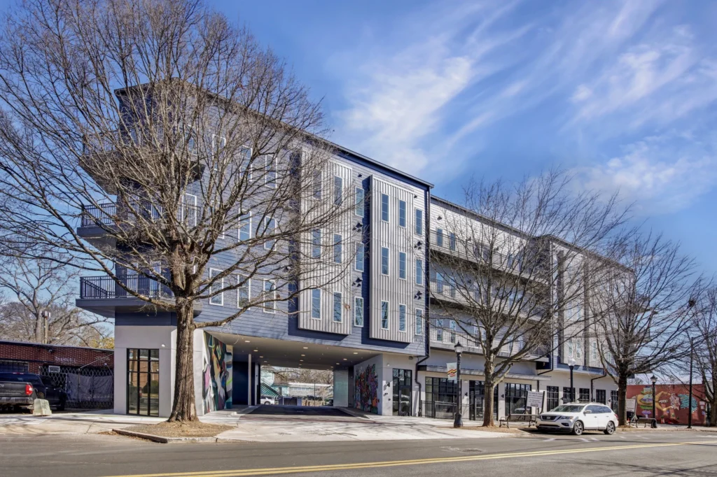 Modern four-story apartment building with large windows, ground-floor retail spaces, bare trees in front, and cars parked along the street on a clear day.