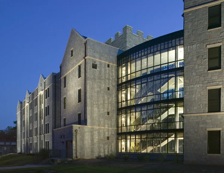 A modern stone building with large glass windows and a visible interior staircase, photographed at dusk with exterior lighting.