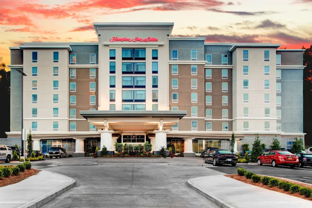 A multi-story Hampton Inn & Suites hotel with a modern exterior, several parked cars in front, and a sunset sky in the background.