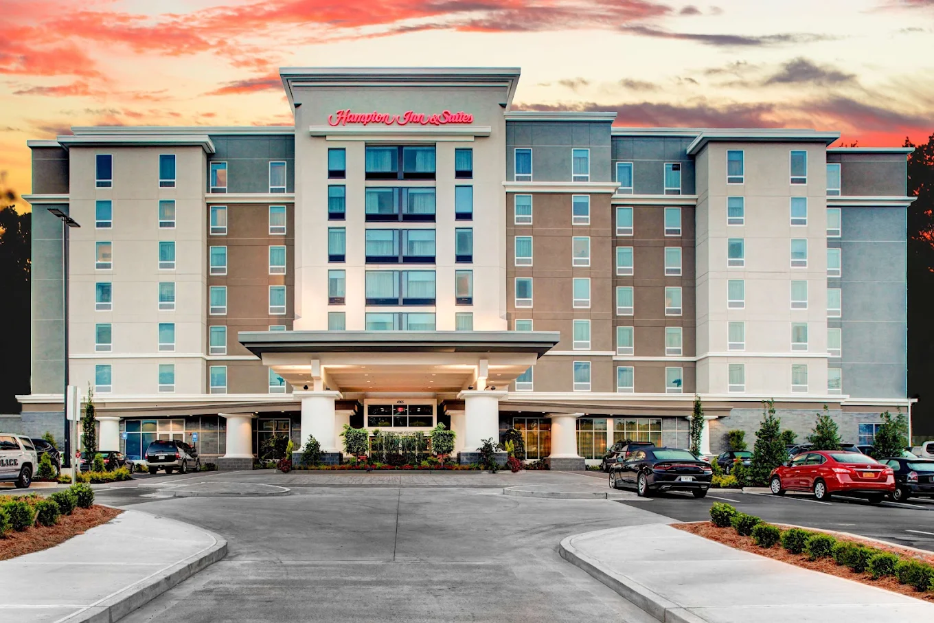 A multi-story Hampton Inn & Suites hotel with a modern exterior, several parked cars in front, and a sunset sky in the background.