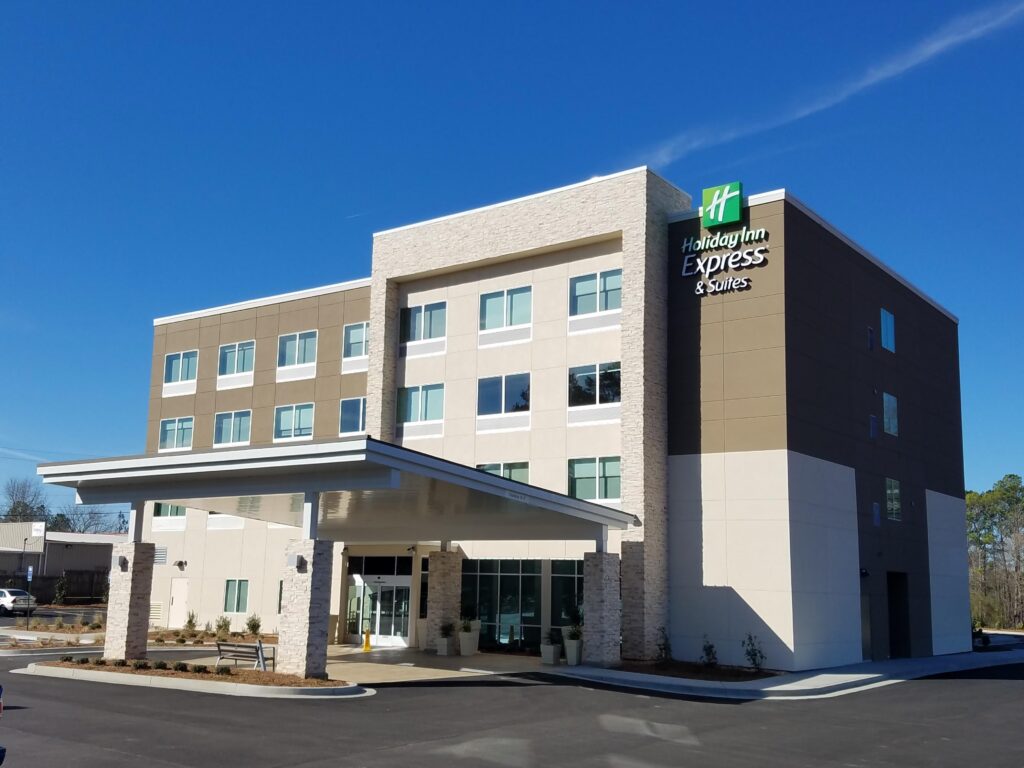 A modern, four-story Holiday Inn Express & Suites hotel with a covered entrance, beige and white exterior, and clear blue sky in the background.