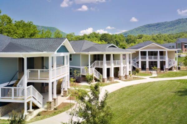 Row of modern, single-story houses with white railings and elevated porches, set on landscaped lawns with hills and trees in the background.