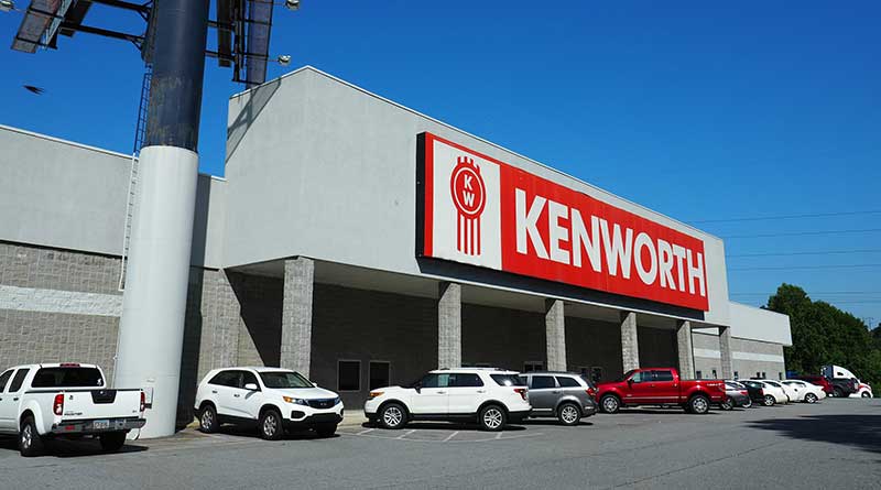 A Kenworth dealership building with a large red and white sign, several white SUVs and trucks parked in front, and a clear blue sky overhead.