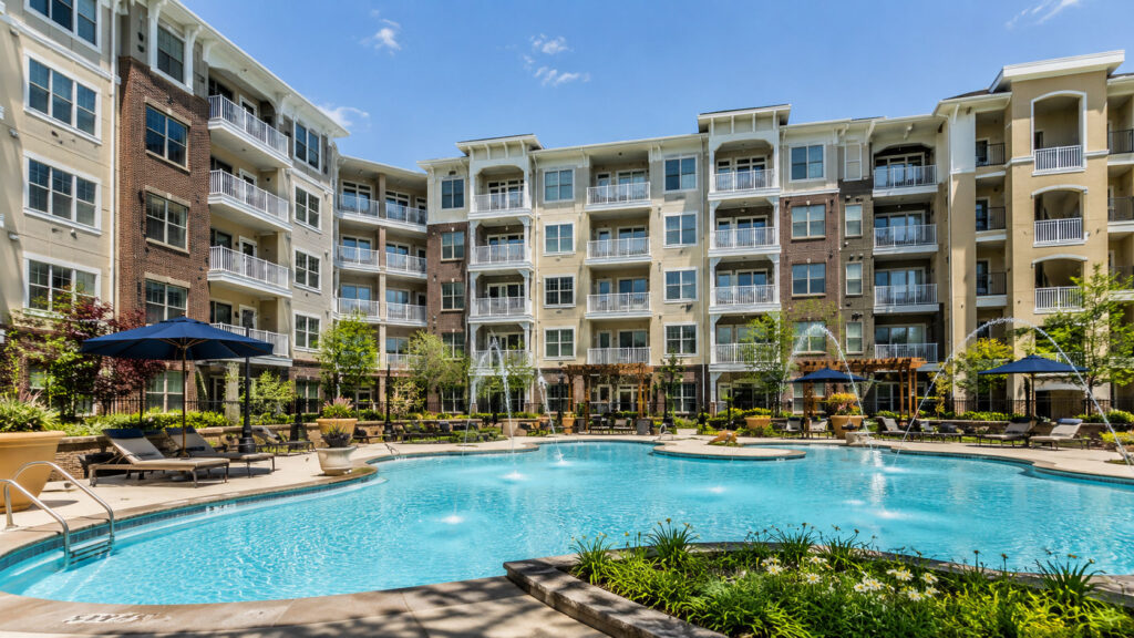 A modern apartment complex with several stories surrounds an outdoor swimming pool, lounge chairs, and umbrellas, under a clear blue sky.