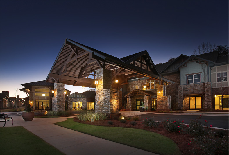 A modern building with a large covered entrance supported by stone pillars, illuminated by outdoor lights, photographed at dusk.