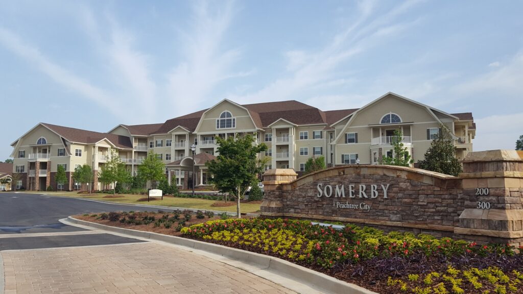 A large, multi-story residential building with balconies, landscaped grounds, and a stone sign reading "SOMERBY Peachtree City" near the entrance.