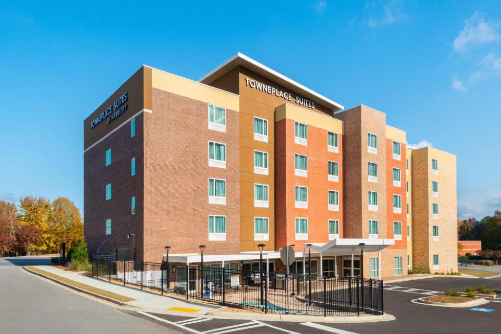 A four-story TownePlace Suites hotel with brick and beige exterior, multiple windows, and a fenced outdoor patio, set against a clear sky and surrounded by parking areas.