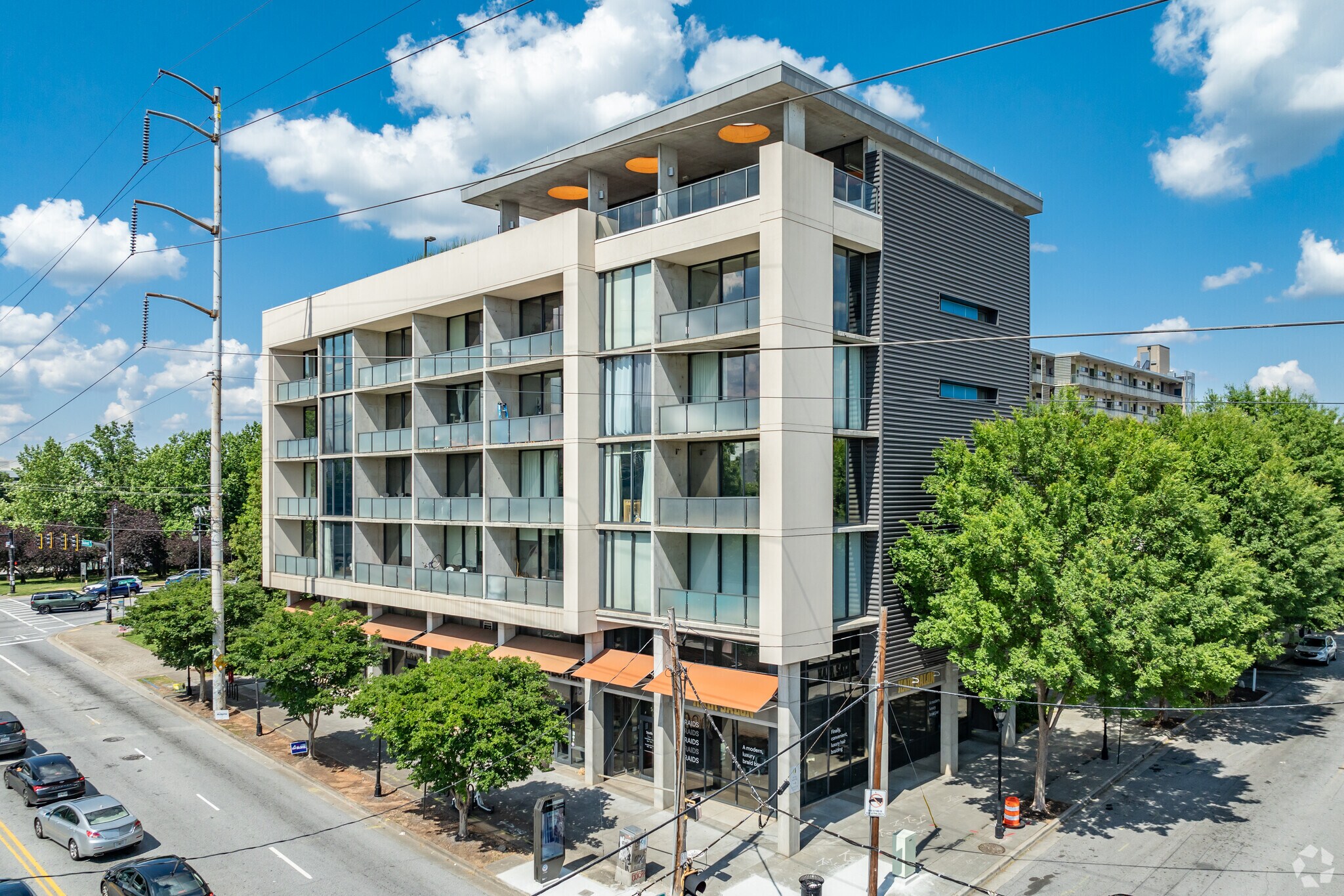 A modern mid-rise apartment building with balconies, large windows, and orange awnings, located at a street corner with trees and parked cars.