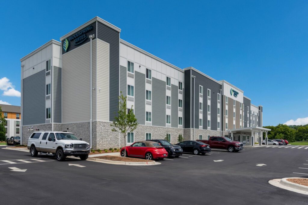 A four-story hotel building with a sign reading "WoodSpring Suites," several parked cars in front, and a clear blue sky overhead.
