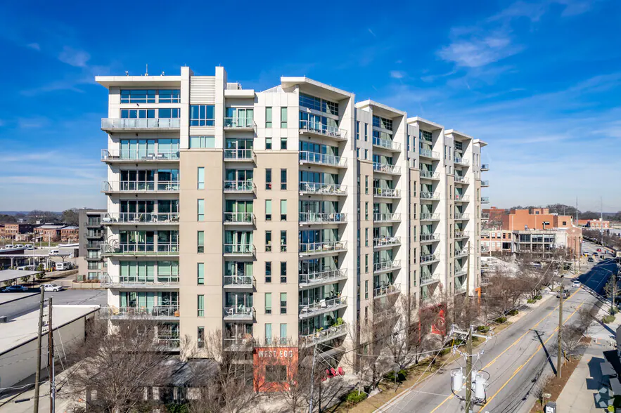 A modern mid-rise apartment building with glass balconies, seen from above on a clear day, with streets and other buildings nearby.