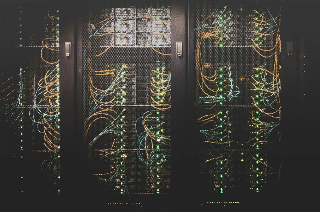 Three black server racks with mesh doors are filled with network cables and blinking green lights, indicating active computer hardware inside a data center.