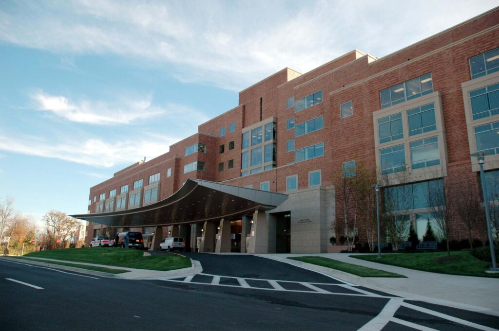 A modern multi-story hospital building with a covered entrance, large windows, and several vehicles parked at the front.