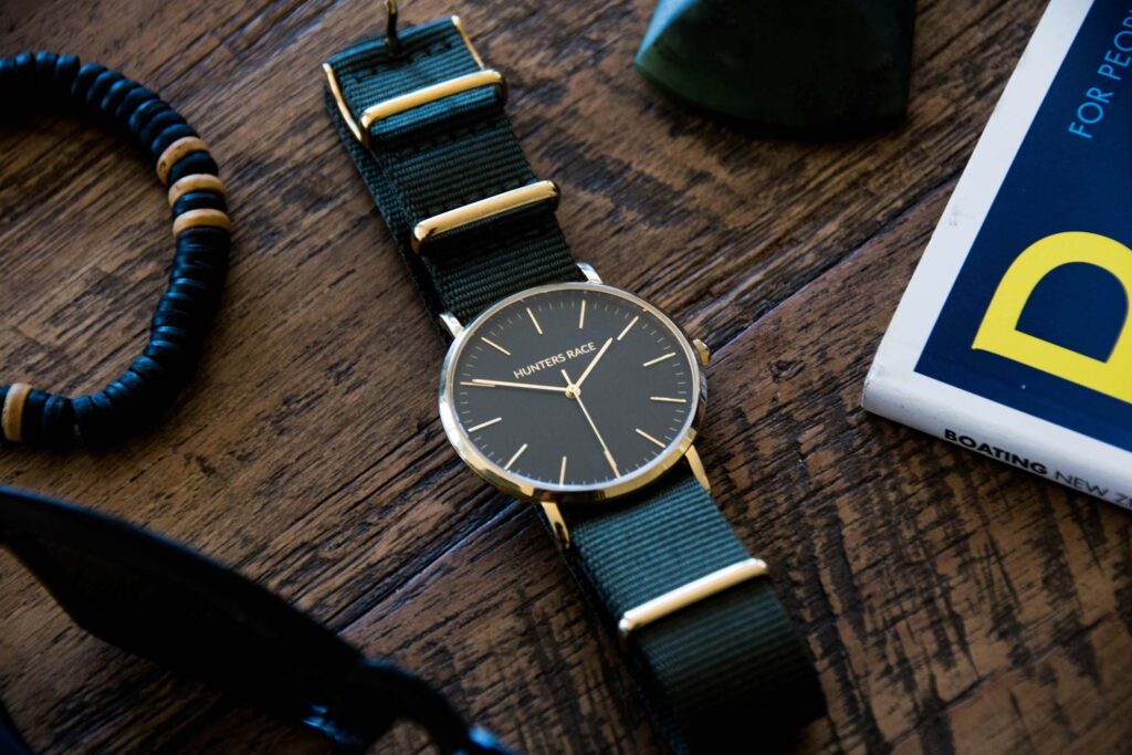 A wristwatch with a green fabric strap and gold accents lies on a wooden surface next to a bracelet, sunglasses, and a partially visible book.