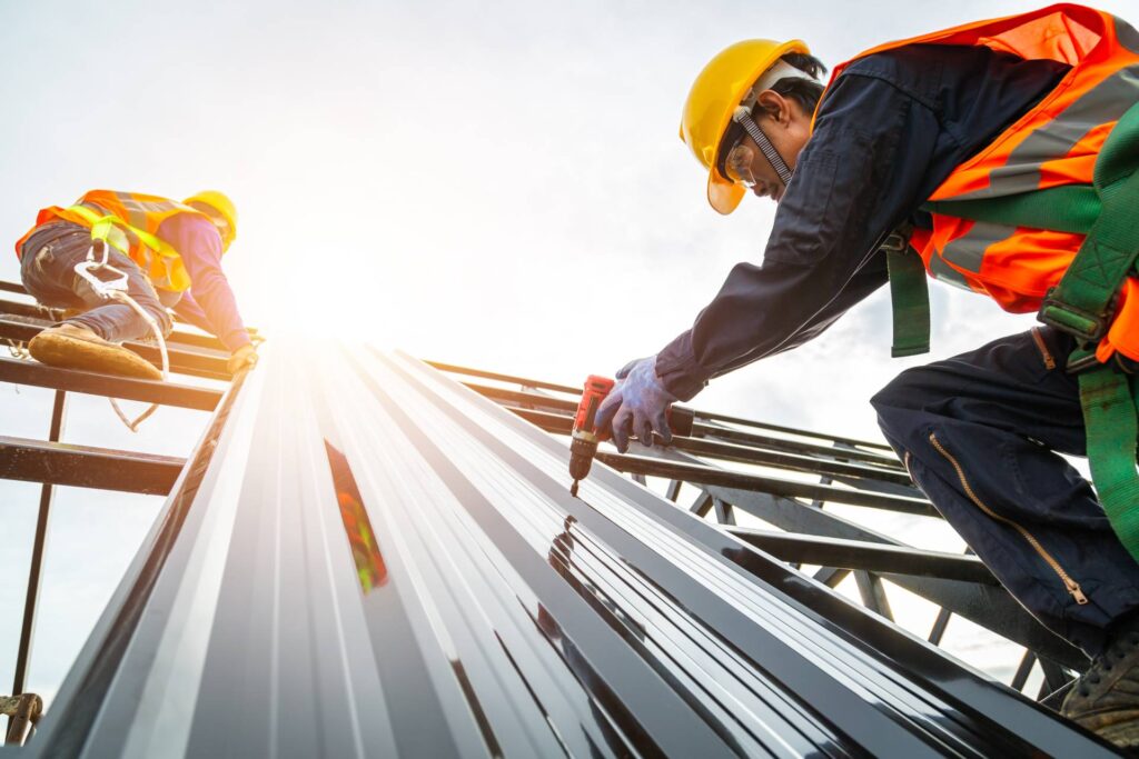 Demonstrating their capabilities, two construction workers in safety gear install metal roofing sheets on a building—one using a power drill, the other climbing the structure—against a backdrop of bright sunlight.