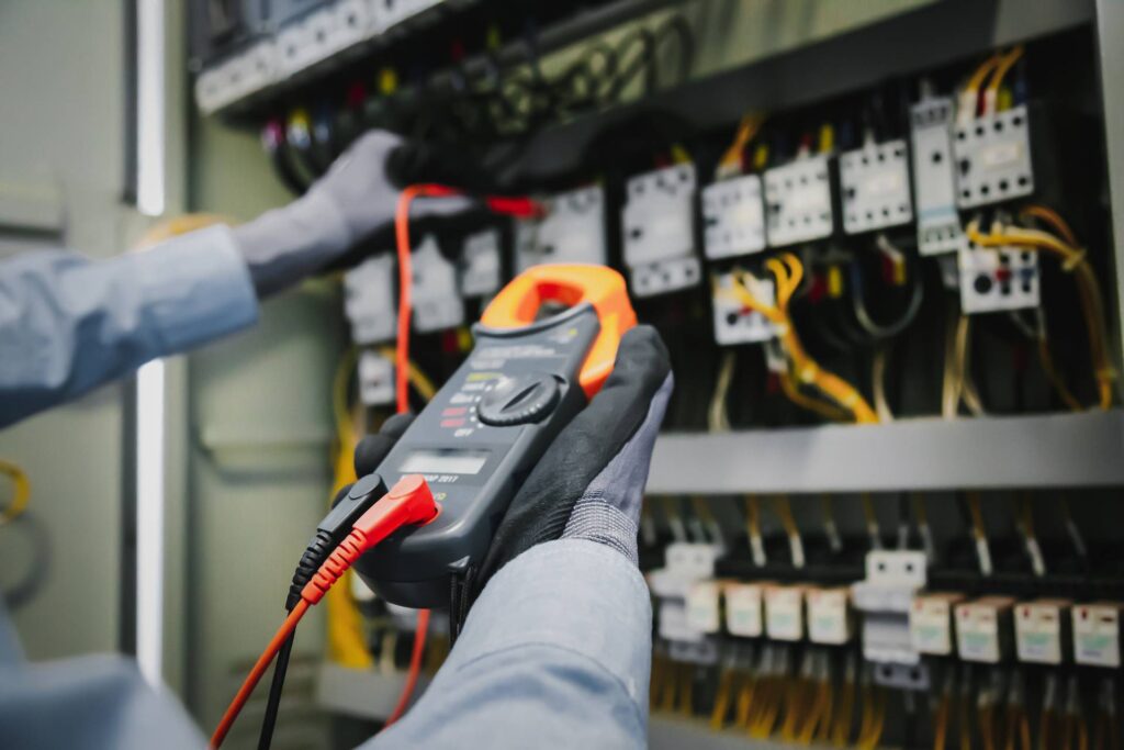 A person wearing gloves uses a digital clamp meter to test the capabilities of electrical connections inside an industrial electrical panel.