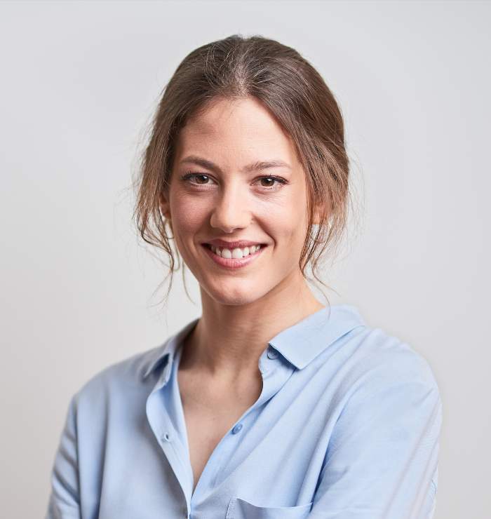 A woman with brown hair in a loose bun, wearing a light blue shirt, smiles at the camera against a plain light background.