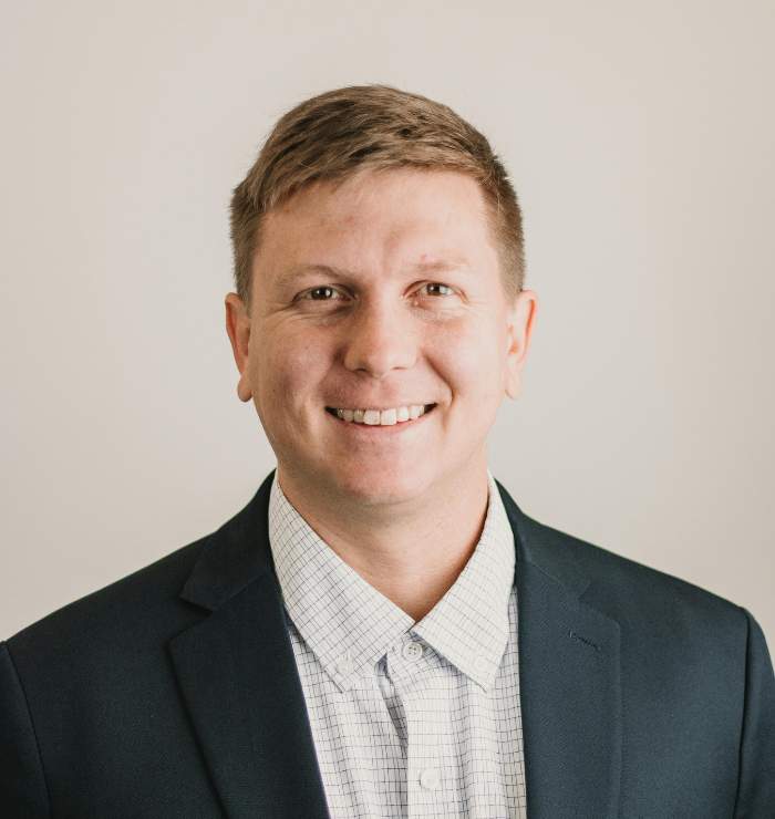 A man in a dark suit jacket and checked dress shirt smiles at the camera against a plain, light-colored background.