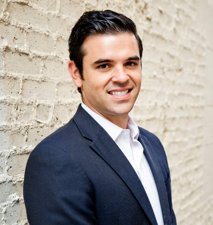A man in a dark suit and white shirt stands in front of a light-colored brick wall, smiling at the camera.
