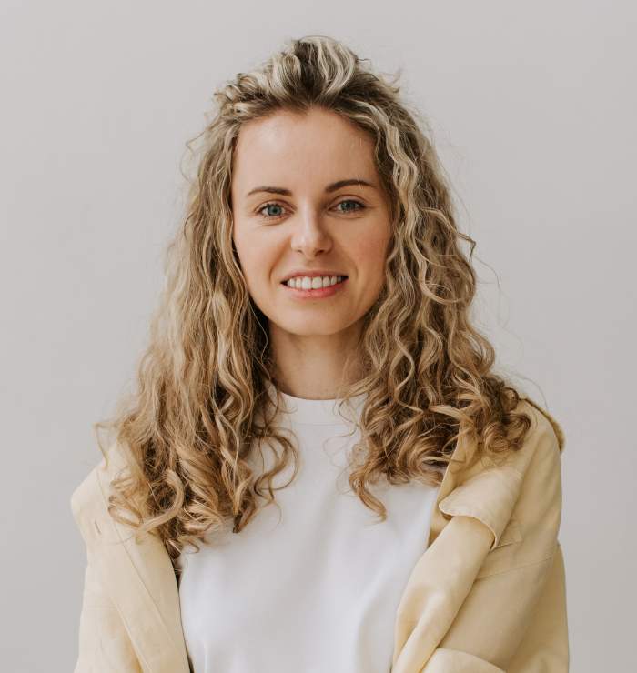 A woman with long curly blonde hair, wearing a white shirt and beige jacket, smiles at the camera against a plain light background.
