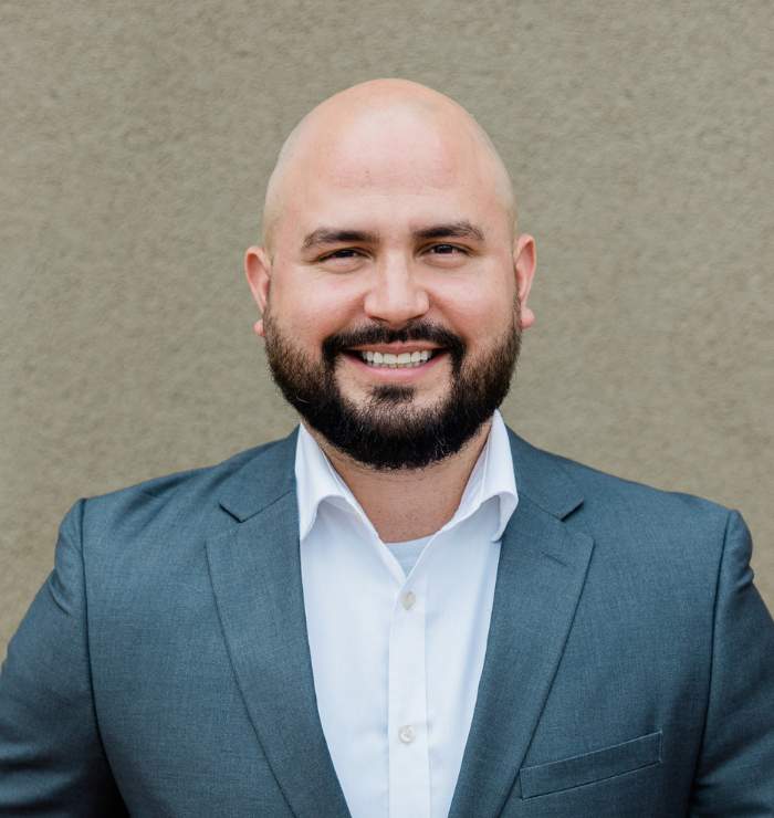 A man with a bald head and beard wearing a gray suit jacket and white shirt stands smiling in front of a plain beige background.