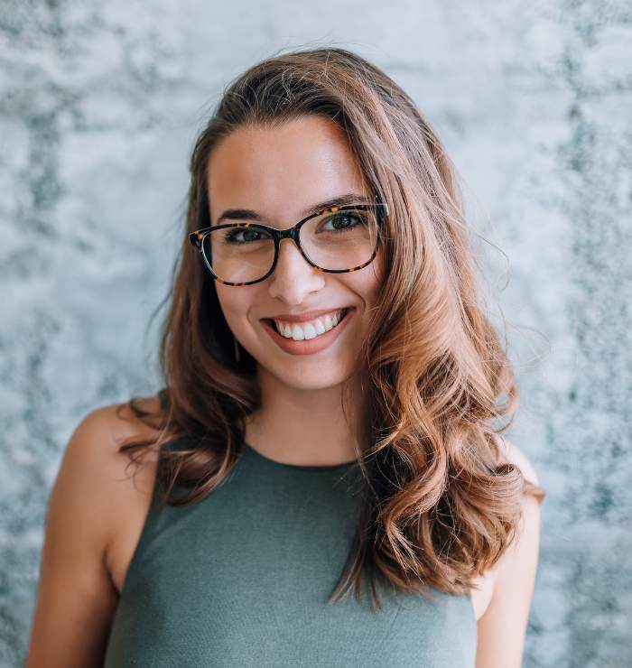 Woman with long brown hair and glasses smiling, wearing a sleeveless green top, standing in front of a textured gray background.